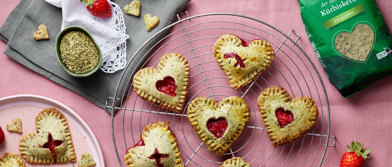 A metal cooling rack holds several heart-shaped pastries with red filling. A strawberry and a green powder bag are nearby.