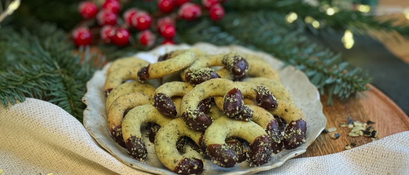 Ein Teller mit Keksen, gefüllt mit Schokolade und Pinienkernen, auf einem Tisch, geschmückt mit Feiertagselementen wie roten Beeren und Lichterketten.