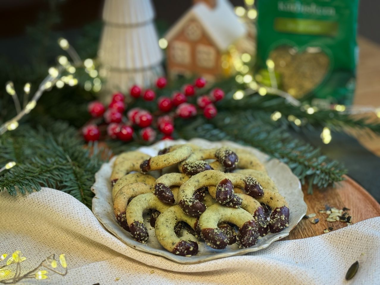 Ein Teller mit grünen Keksen und Schokolade auf einem Tisch, dekoriert mit roten Beeren und Lichtern, und einem Weihnachtshaus im Hintergrund.