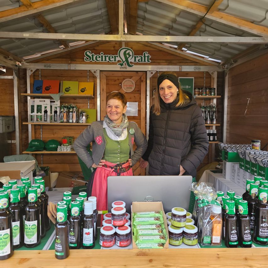 Two women stand behind a counter in a shop, smiling. They are surrounded by various products, including bottles of sauce and jars of jam. A laptop is on the counter. The shop has a wooden interior.