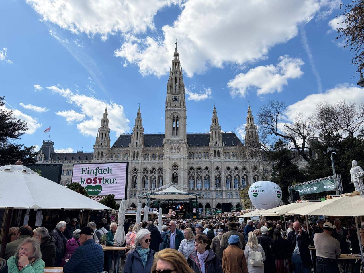 A large crowd gathers at an outdoor event in front of Vienna's Rathaus, with a sign advertising a cost-free tour.