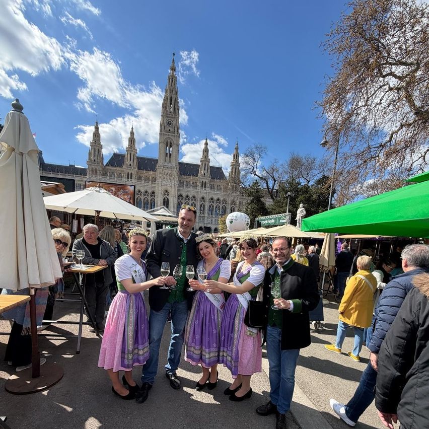 A group of people dressed in traditional attire, including two women in dirndls, are posing for a photo with wine glasses in hand at an outdoor event. In the background is a large building with a clock tower.