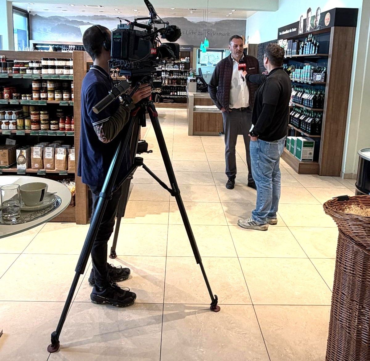 Ein Mann mit einer Kamera auf einem Stativ führt ein Interview mit zwei Männern in einem Geschäft.