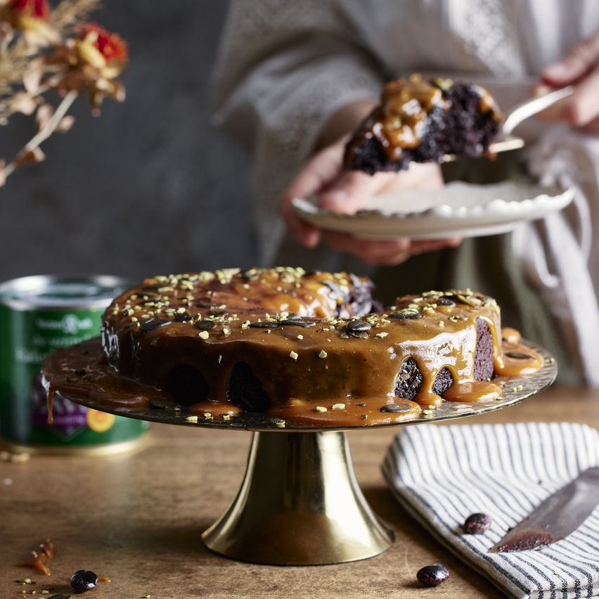 Eine Person hält ein Stück Schokoladenkuchen auf einem silbernen Kuchenständer. Der Kuchen ist mit Karamell und Nüssen garniert, und ein Messer liegt in der Nähe.