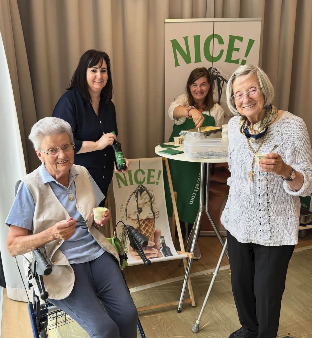 Four women stand together in a room, two of them holding cups. One woman is in a wheelchair. Behind them, a table holds a bottle and a sign that reads 'NICE!'.