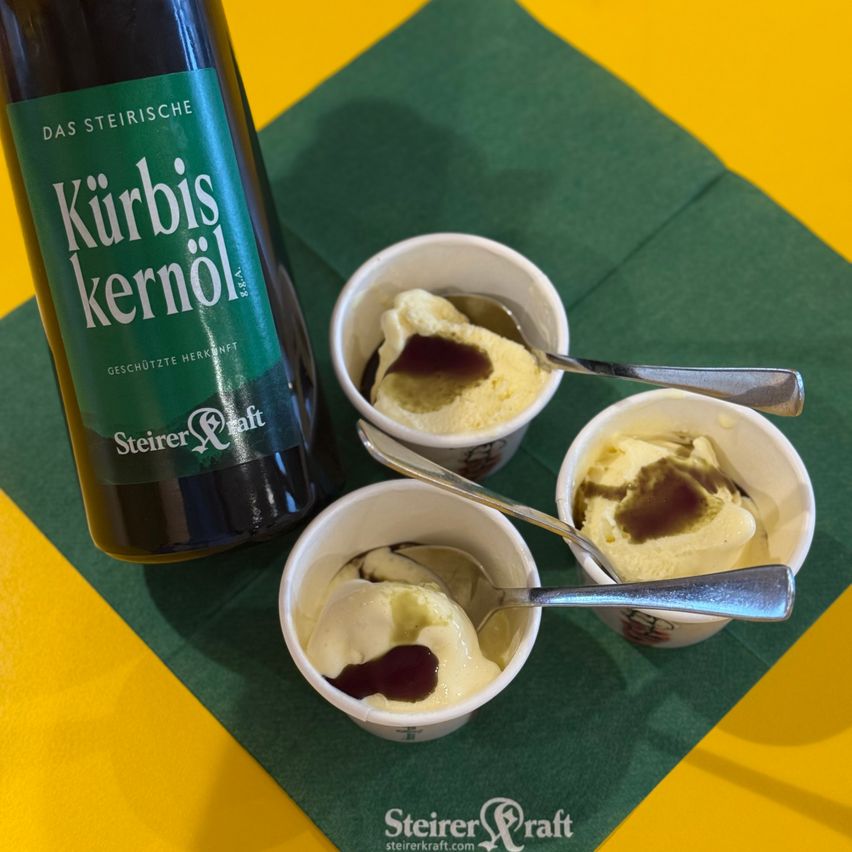 Three small cups of ice cream with a drizzle of red sauce and a bottle of Kürbis kernöl on a green cloth.