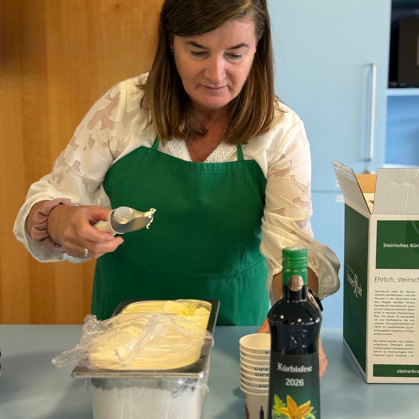 A woman in a green apron is scooping yellow cream into a container on a table. A bottle of 'Kürbisfest 2026' and a box with the same label are nearby.