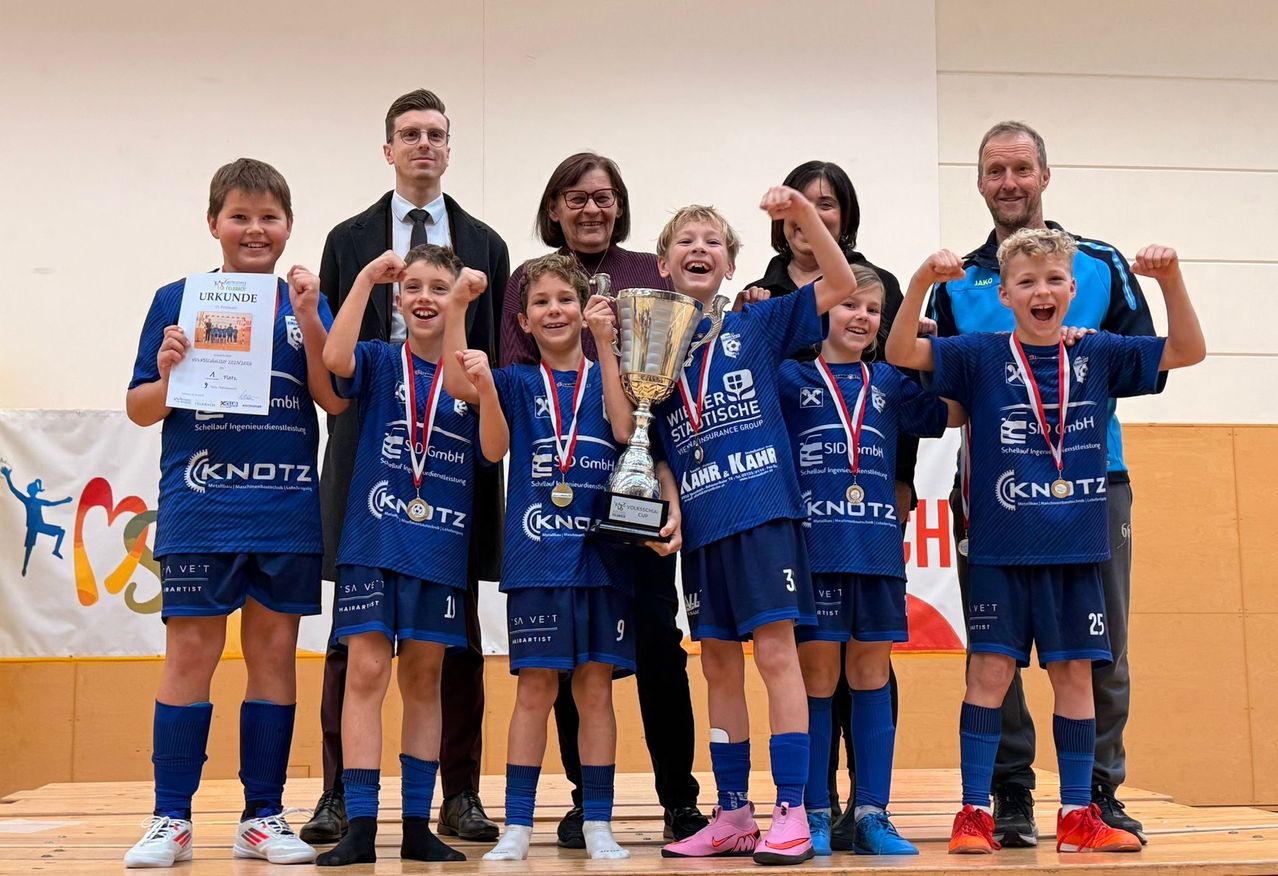 Eine Gruppe junger Athleten in blauen Uniformen posiert mit ihrem Trainer, hält einen Pokal und Urkunden in der Hand, während Erwachsene hinter ihnen lächeln.
