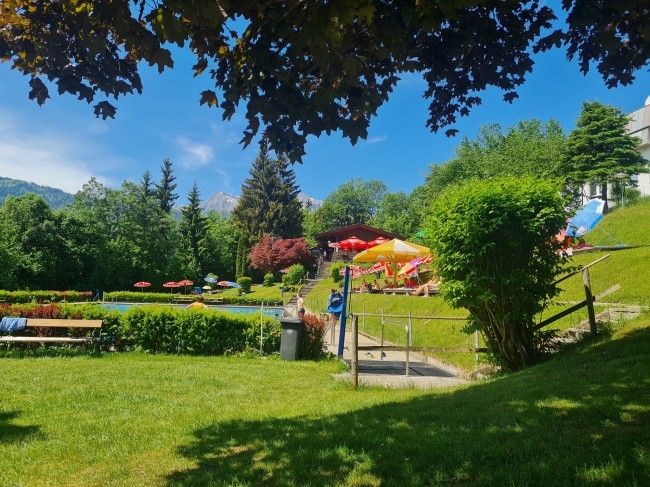 Ein Ferienort mit Schwimmbad, bunten Sonnenschirmen und einem baumbestandenen Grashügel.