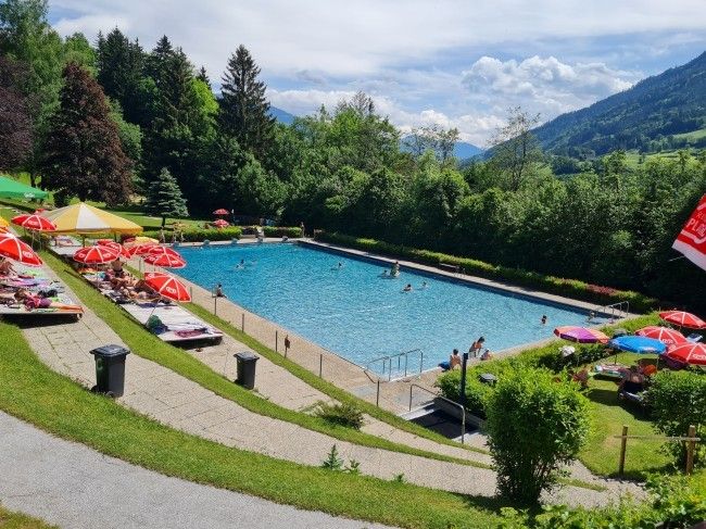 Ein Freibad umgeben von viel Grün, mit mehreren schwimmenden und unter bunten Sonnenschirmen liegenden Personen. Berge und Bäume sind im Hintergrund zu sehen.