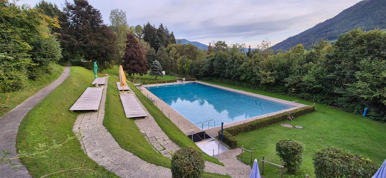 Eine Vogelperspektive eines großen Außenschwimmbeckens, umgeben von üppigem Grün und Bergen im Hintergrund.