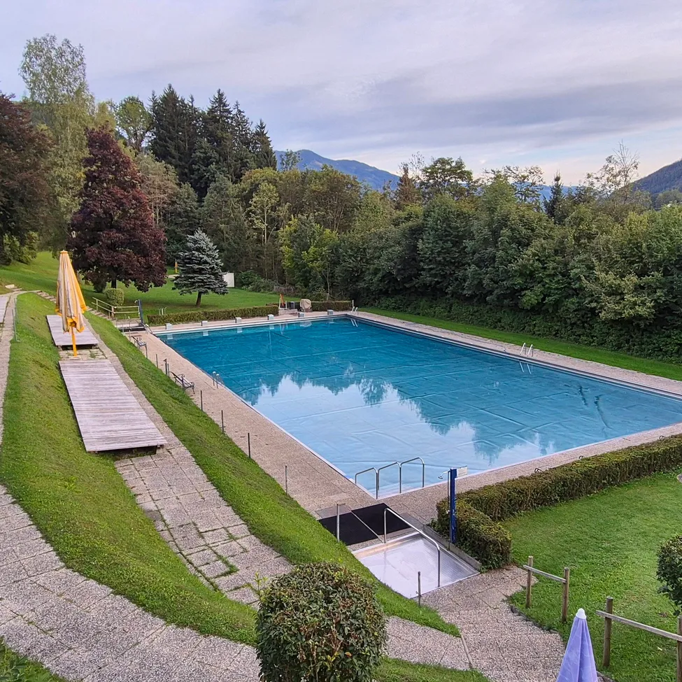 Eine Vogelperspektive eines großen Außenschwimmbeckens, umgeben von üppigem Grün und Bergen im Hintergrund.