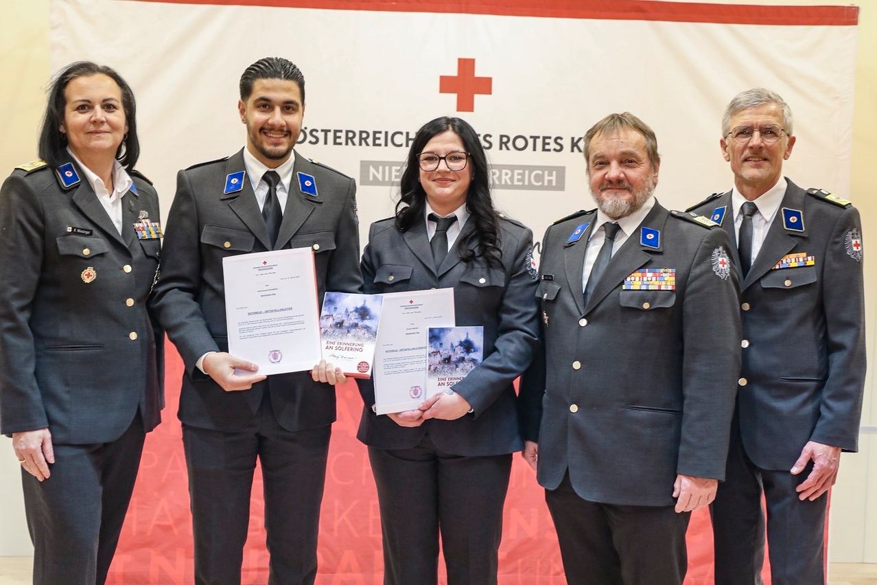 Vier uniformierte Personen stehen vor einem Hintergrund mit einem roten Kreuz. Zwei Männer und eine Frau halten Urkunden in den Händen. Die Frau trägt eine Brille. Der Mann rechts hat einen Bart und Medaillen auf der Brust.