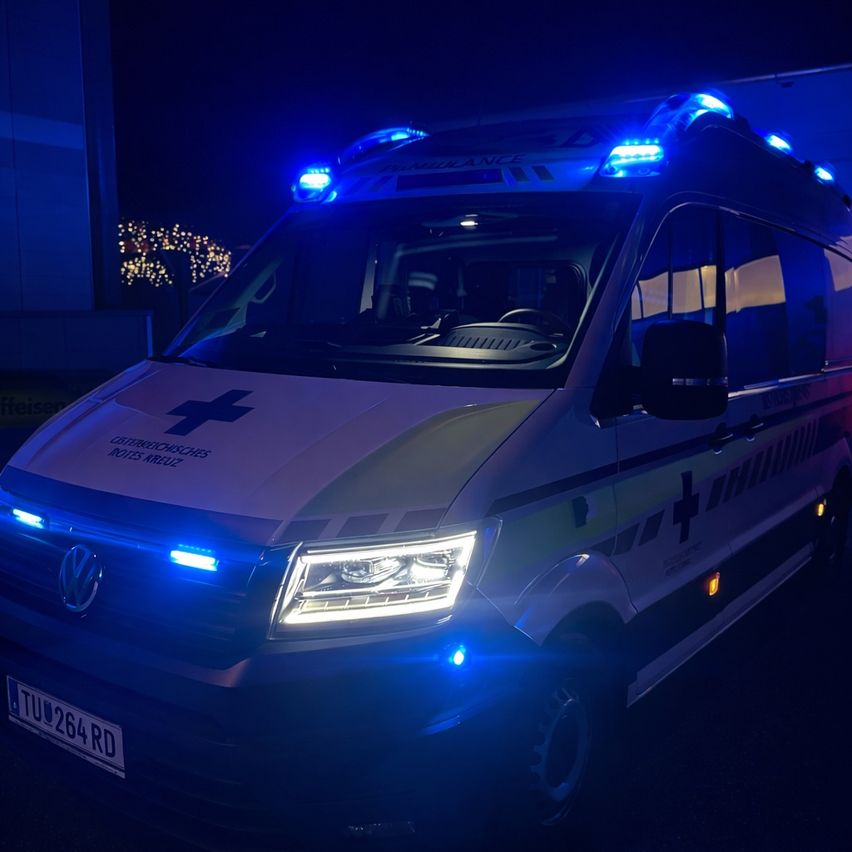 An ambulance with emergency lights on is parked at night. The front of the vehicle has a red cross and the word 'Ambulance'. The license plate reads 'TU0264RD.'