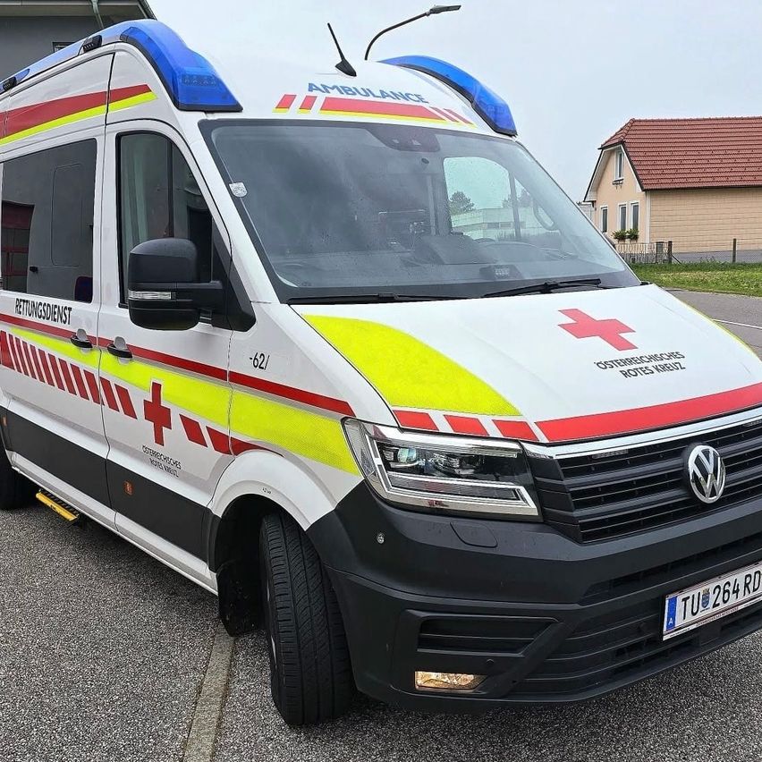 A white ambulance with red and yellow stripes is parked on a street with houses in the background. It has a red cross on the side and a license plate.