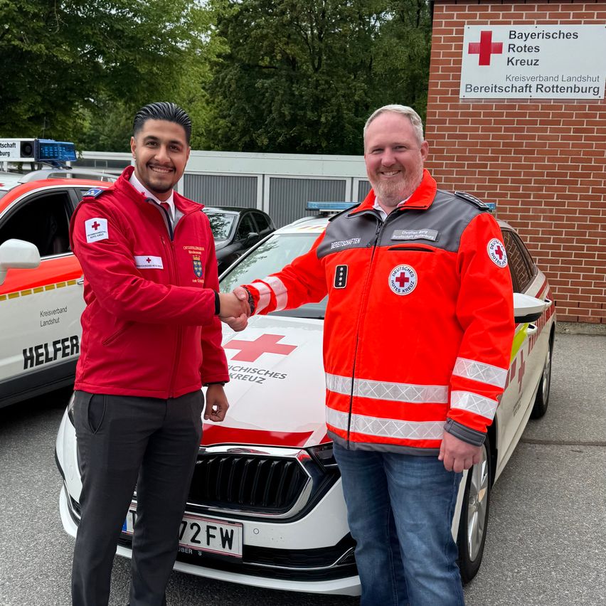 Two men in red and white safety jackets shake hands in front of a white car with a red cross logo. Behind them are two white emergency vehicles. A sign reads Bayerisches Rotes Kreuz Kreisverband Rottenburg.
