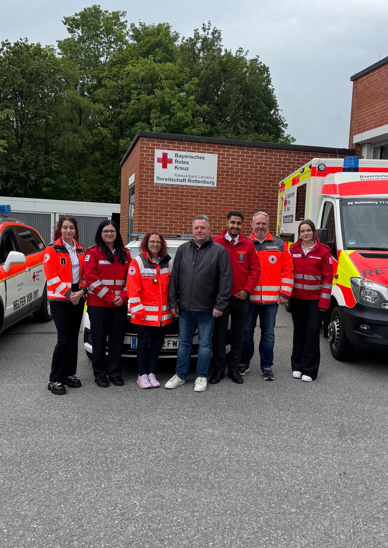 A group of people in orange vests stand in front of a brick building, likely a Red Cross facility. An ambulance is parked beside them.