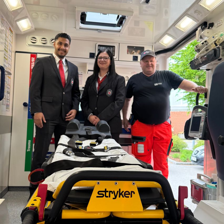 Three people are standing inside an ambulance with a Stryker stretcher. One person is wearing a red tie and the other two are in uniforms.