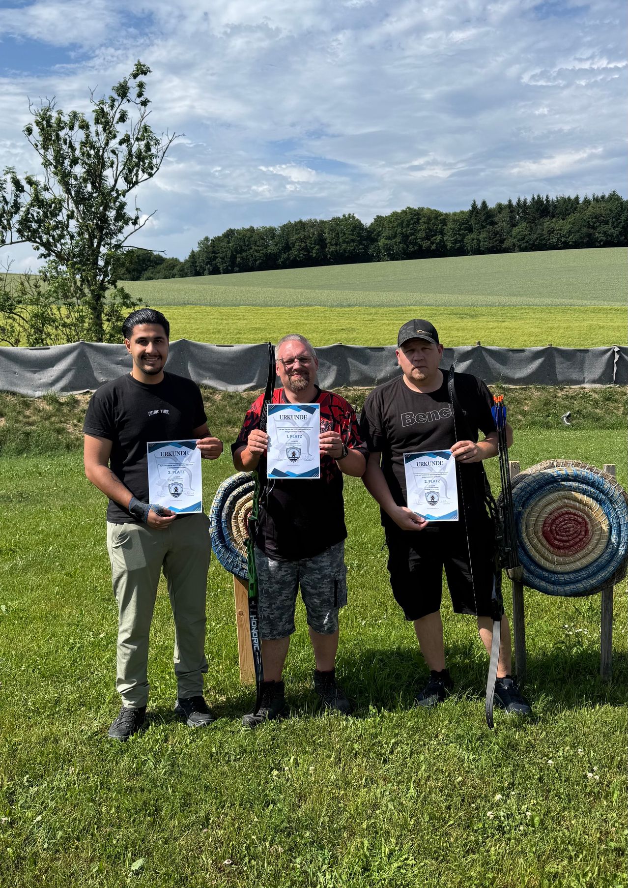 Three men stand in a field, holding certificates and bows. They are smiling and posing for a photo. Behind them is a grassy field and a tree. The sky is partly cloudy.