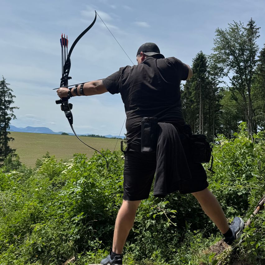 A man is outdoors, holding a bow and aiming at a target with three arrows. He wears black clothes and shoes. Behind him, a field and trees are visible.