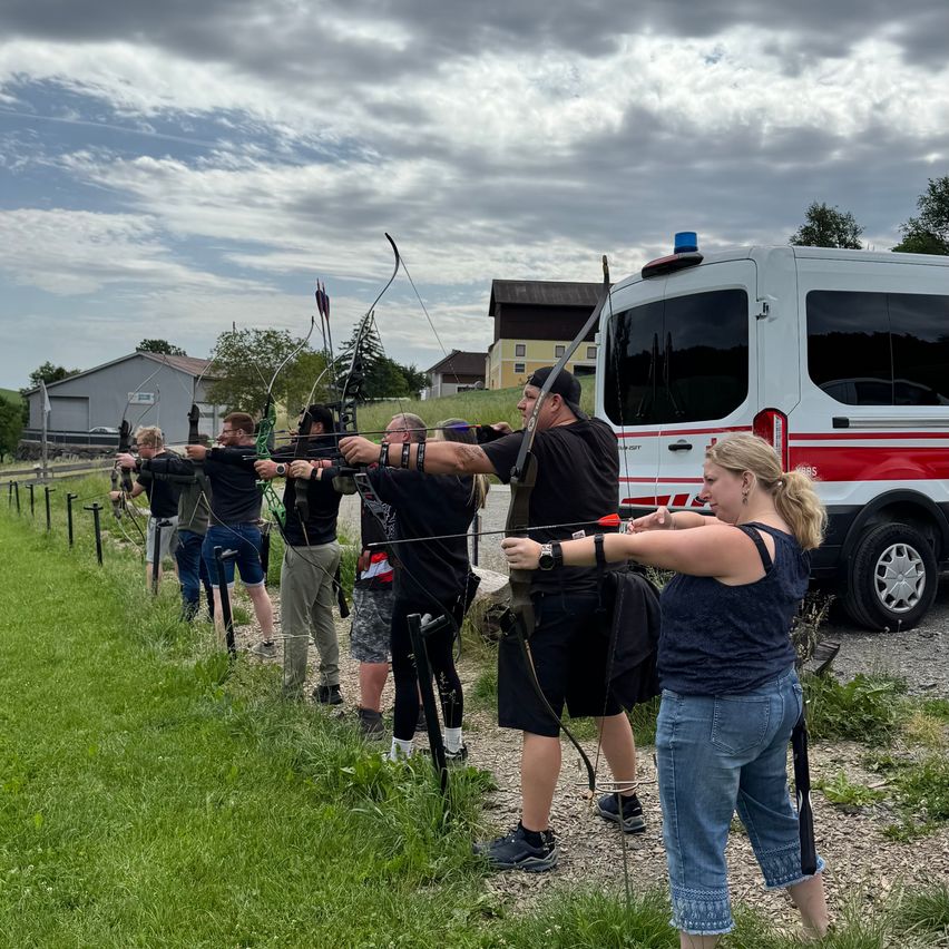 A group of people practices archery in a grassy area, with a white van parked behind them on the side. They are all wearing hats and some are wearing bracelets.
