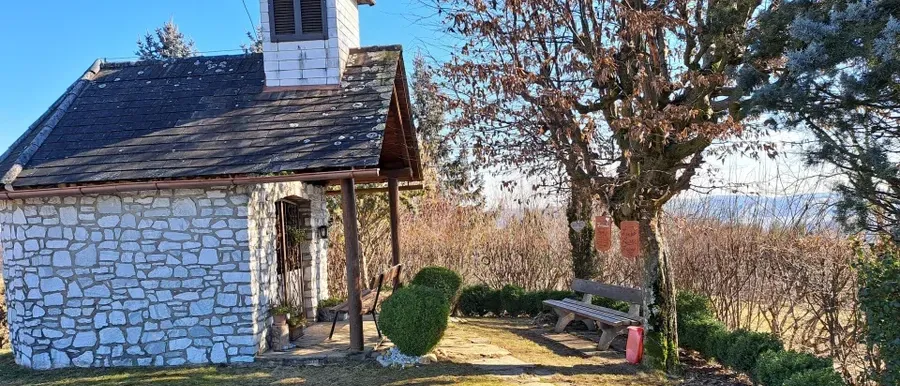 Eine kleine Kapelle aus Stein mit einem hölzernen Vorbau, einer Bank und einem Baum steht in einem Garten.