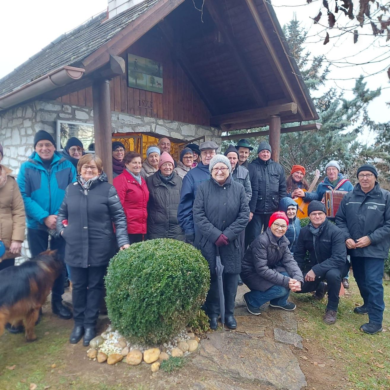 Eine Gruppe von Menschen in Winterkleidung, die vor einem rustikalen Haus mit Steinmauer und Dach lächelnd für ein Foto posieren. Ein Busch und ein Hund sind ebenfalls vorhanden.