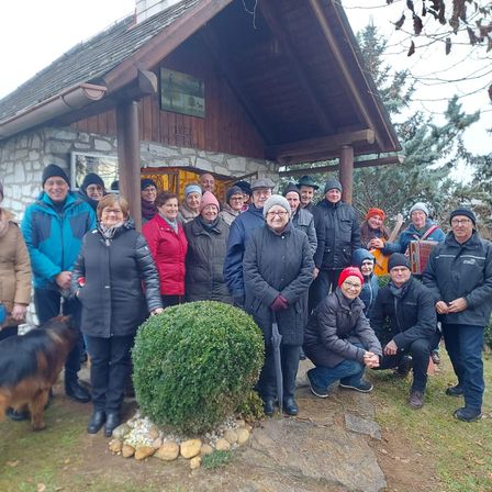 Eine Gruppe von Menschen in Winterkleidung, die vor einem rustikalen Haus mit Steinmauer und Dach lächelnd für ein Foto posieren. Ein Busch und ein Hund sind ebenfalls vorhanden.