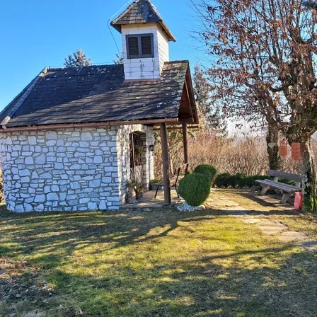 Eine kleine Steinkapelle mit einem kleinen Turm. Eine Bank und ein Baum sind im Hof. Ein Busch steht vor dem Haus.