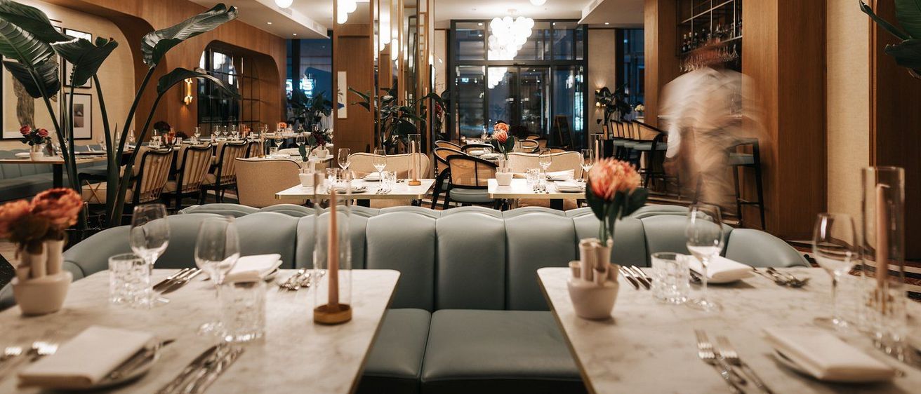 Ein elegantes Restaurant mit Holzverkleidungen, modernen Möbeln und großen Fenstern. Die Tische sind mit Blumen, Weingläsern und Besteck gedeckt. Eine Person geht im Hintergrund.