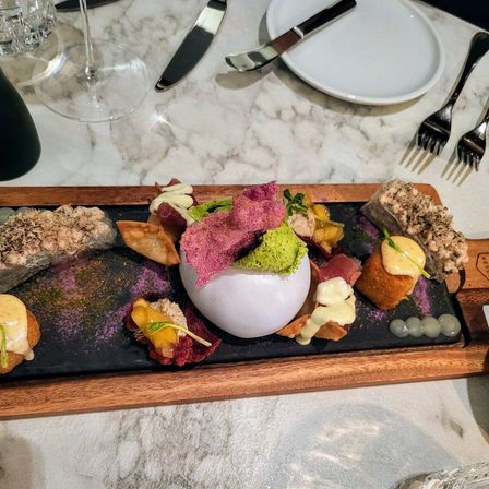 Ein schwarzes Tablett mit verschiedenen Speisen, darunter eine weiße Schüssel mit rosa Füllung, garniert mit grünen und rosa Elementen. Im Hintergrund befinden sich ein Weinglas, eine Flasche und Besteck.