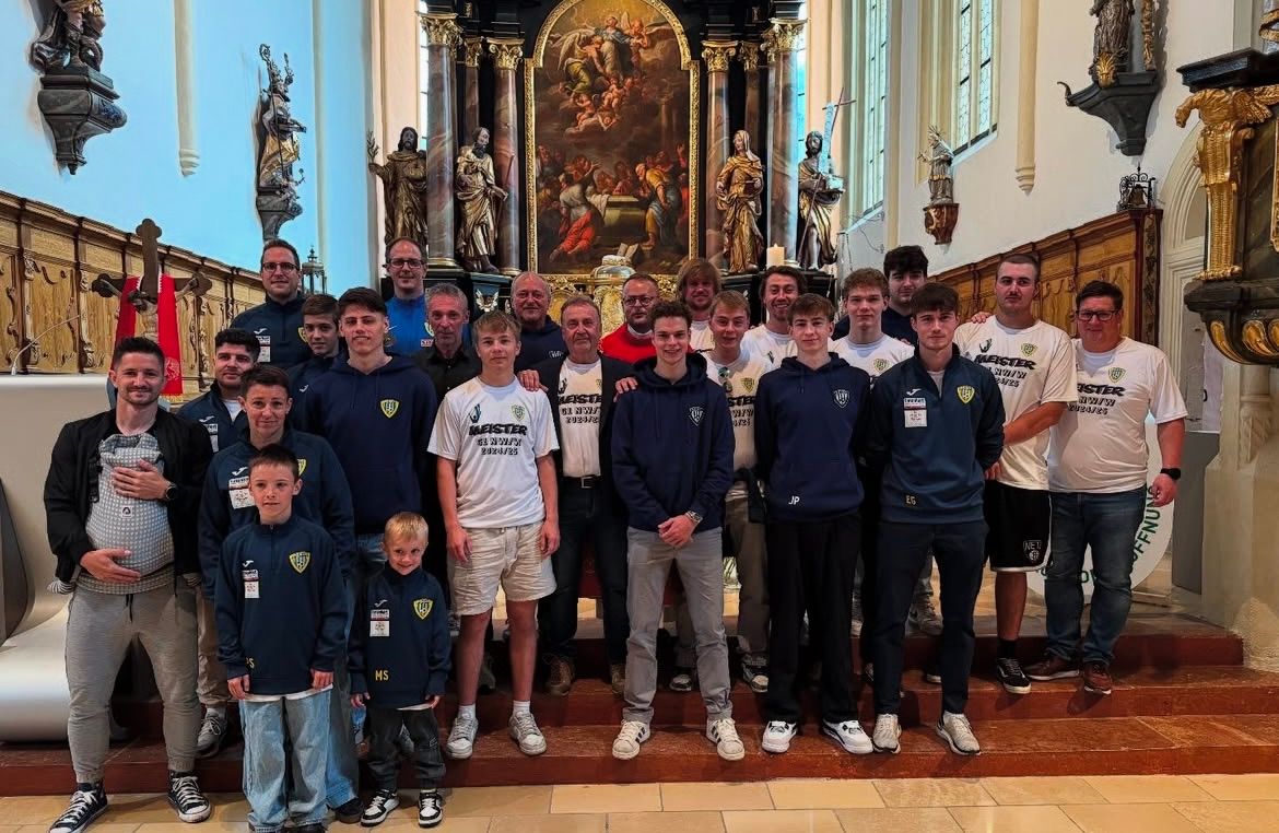 Eine Gruppe von Männern und Jungen in Sportkleidung, lächelnd, steht vor einem Kirchenaltar, mit Statuen und Gemälden im Hintergrund.