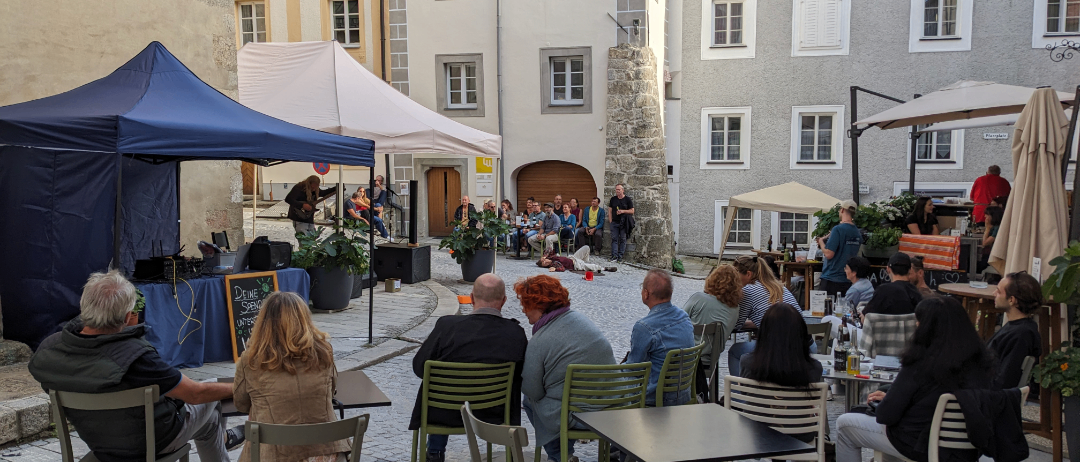 A group of people sit around tables in a patio area, with tents and a small stage setup for a performance. The buildings in the background are white with multiple windows.