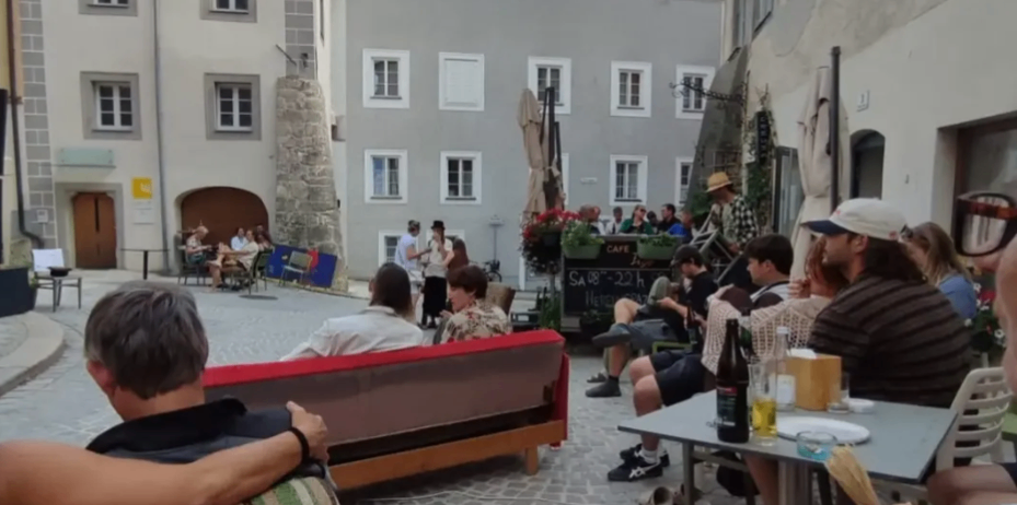 Eine Gruppe von Menschen sitzt auf Bänken und Stühlen um einen kleinen Platz, einige stehen und unterhalten sich. Im Hintergrund befinden sich Gebäude mit vielen Fenstern.