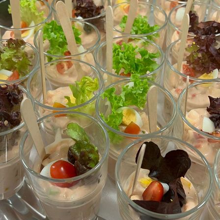 Eine Nahaufnahme mehrerer transparenter Gläser, gefüllt mit einem rosafarbenen Gericht, das mit Salat, Tomaten und Eiern belegt ist. In jedes Glas ist ein Holzstäbchen eingefügt.