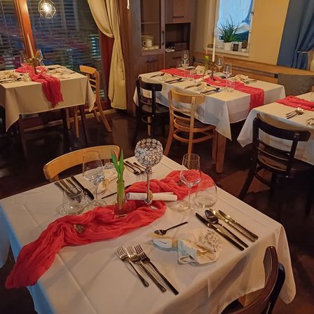 Ein Restaurant mit mehreren Tischen, die mit weißen Tischdecken, Besteck und dekorativen Servietten gedeckt sind. Ein Tisch hat einen Mittelpunkt mit einem roten Tuch und einer klaren Glasvase.