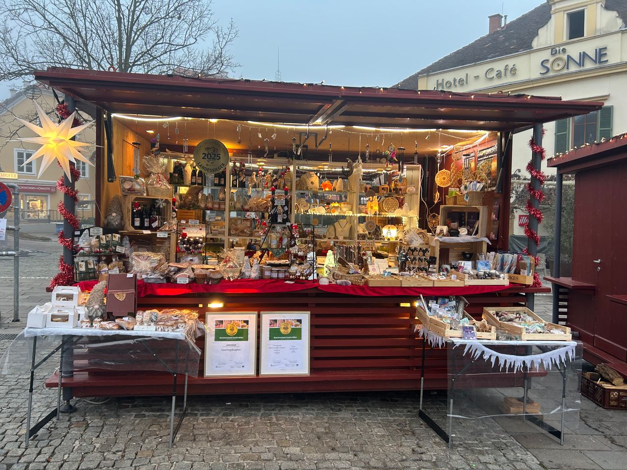 Ein Marktstand mit festlicher Gestaltung, beleuchtet mit Lichtern, zeigt eine Vielzahl von Produkten, darunter Weinflaschen, Geschenkverpackungen und handgefertigte Kunst. Zertifikate sind vorne ausgestellt.