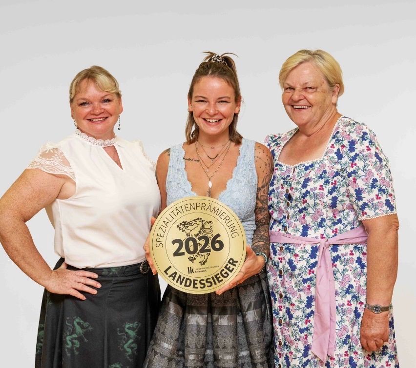 Drei Frauen stehen lächelnd zusammen, eine hält eine goldene Medaille mit der Aufschrift '2026 Landessieger'. Die Frau in der Mitte trägt ein ärmelloses Kleid und Halsketten. Die anderen tragen weiße und blumige Kleider.
