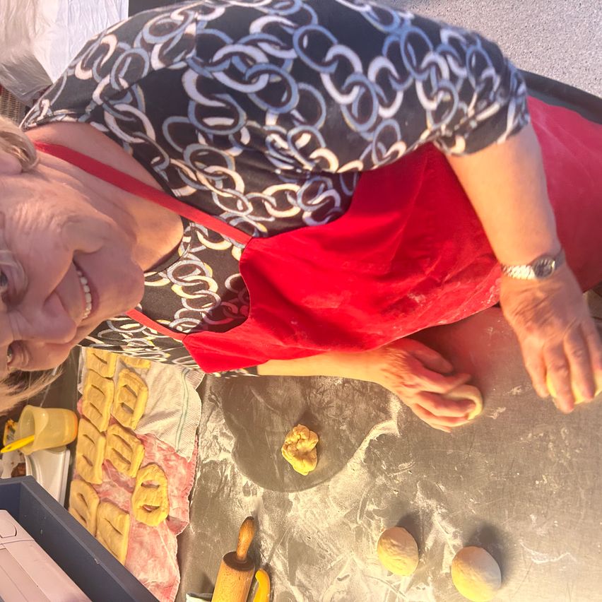 Eine ältere Frau kniet und lächelt, während sie Teig auf einer Arbeitsplatte vorbereitet, mit einem Nudelholz und drei Teigkugeln in der Nähe. Sie trägt ein rotes Schürzen und hat mehrere geformte Teigstücke auf einem Tuch hinter ihr.