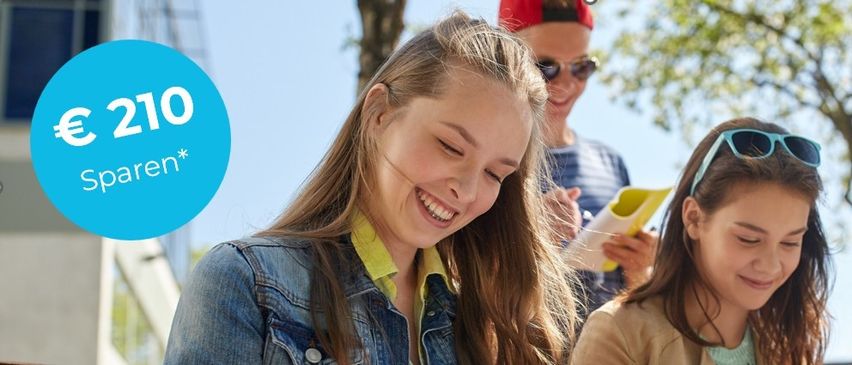 Zwei junge Mädchen lächeln, eines trägt eine Jeansjacke, das andere ein beiges Top. Eine Person hinter ihnen hält ein gelbes Objekt. Sie scheinen sich in einem Park zu befinden.