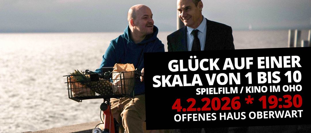 Zwei lächelnde Männer neben einem Fahrrad, einer hält einen Korb mit einer Ananas, der andere ist in einem Anzug. Vor ihnen steht ein Plakat: 'Glück auf Skala von Spielfilm 4.2.2026 Offenes Haus'.