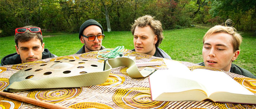Drei Männer in einem Park, einer mit einer Mütze, ein anderer mit Brille, schauen über einen Tisch mit einem Buch, einem Holzstock und einer Filmrolle.