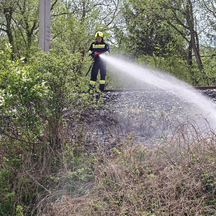 Ein Feuerwehrmann in Schutzausrüstung steht auf einem Bahngleis und sprüht Wasser, um ein Feuer zu löschen.