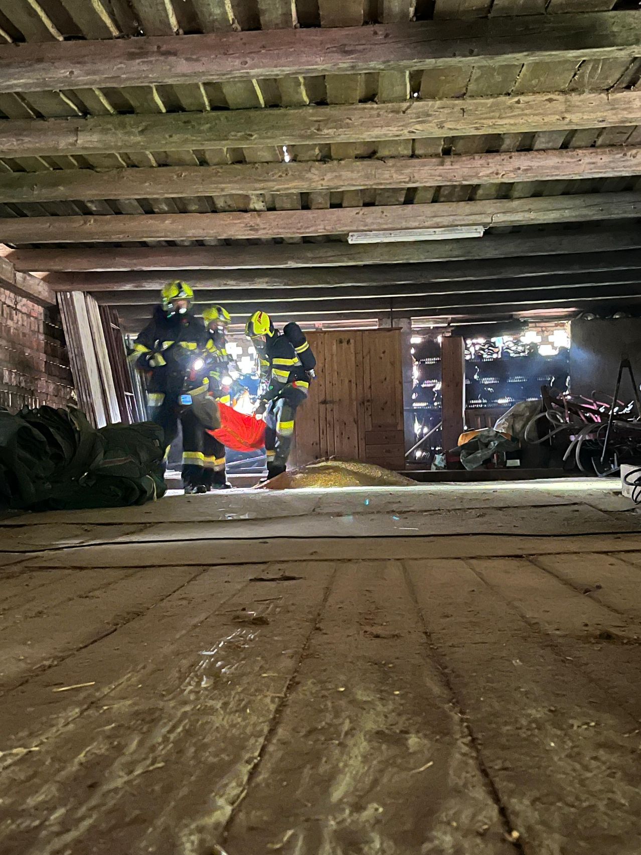 Three firefighters are in a tunnel with a yellow object on the floor. They wear helmets and carry backpacks. Wooden beams and a wooden door are on the right.