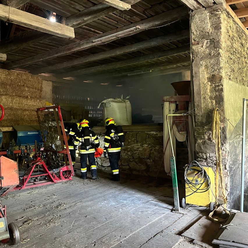 Three firefighters in protective gear stand in a smoke-filled barn. Equipment and tools are scattered around. A large stone wall is in the background, and a red machine is on the left.