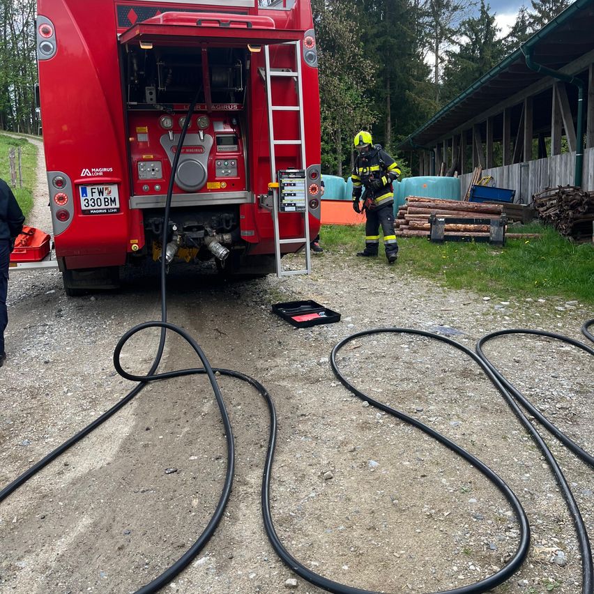 A firefighter is preparing a fire truck, with hoses laid out on the ground, in a rural area with trees and a wooden structure.