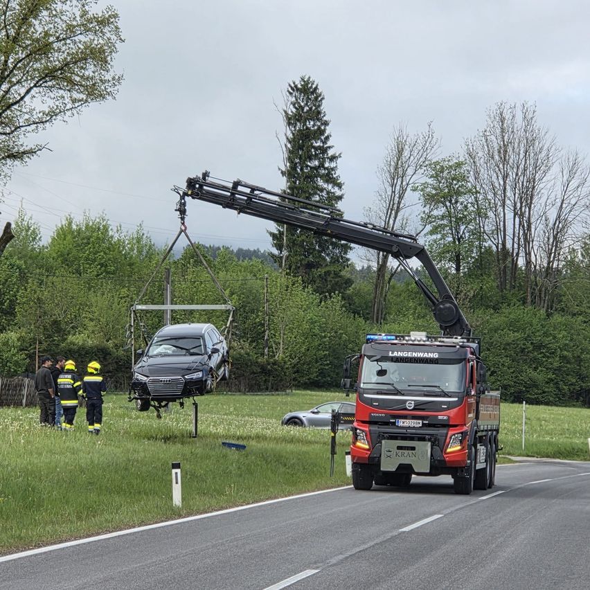 A crane is lifting a damaged black car off the roadside. Firefighters observe the scene. A red truck drives by.
