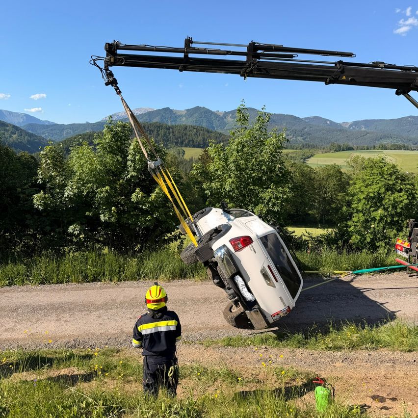 Ein weisses SUV wird von einem Kran auf einer Kiesstrasse angehoben, ein Feuerwehrmann beobachtet. Im Hintergrund sind grüne Felder und Berge zu sehen.