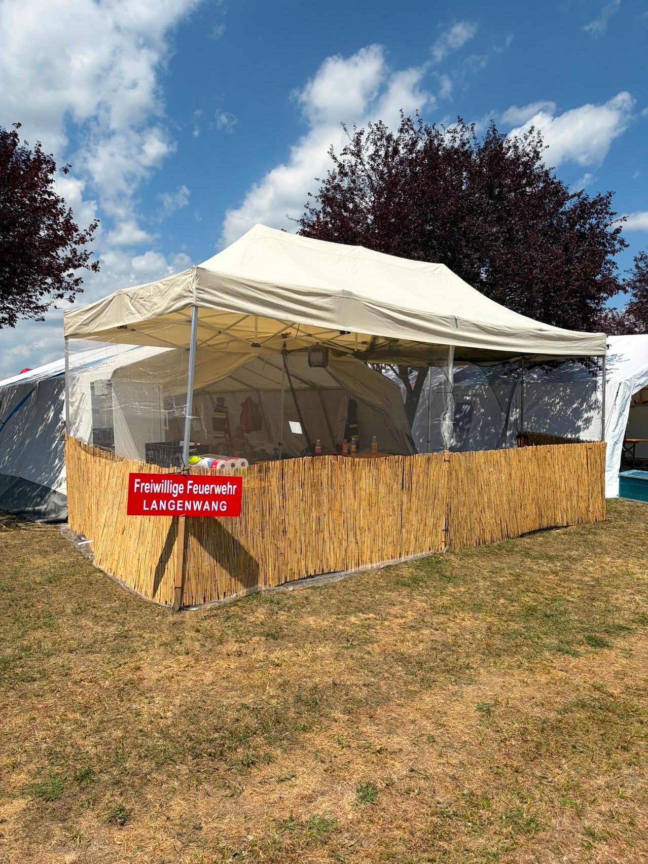Ein weißes Zelt mit einem roten Schild, das 'Freiwillige Feuerwehr LANGENWANG' steht auf einem Rasenfeld mit Bäumen und einem weiteren Zelt im Hintergrund.