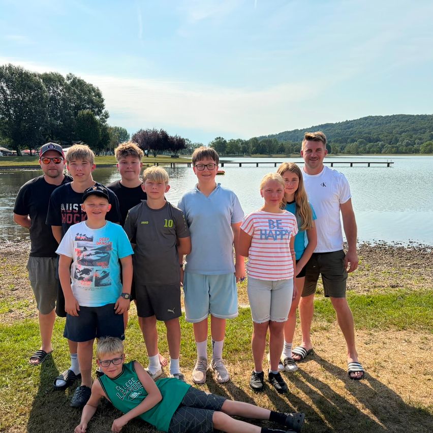Eine Gruppe junger Menschen und Erwachsener posiert für ein Foto am See, mit einer Brücke und Hügeln im Hintergrund.